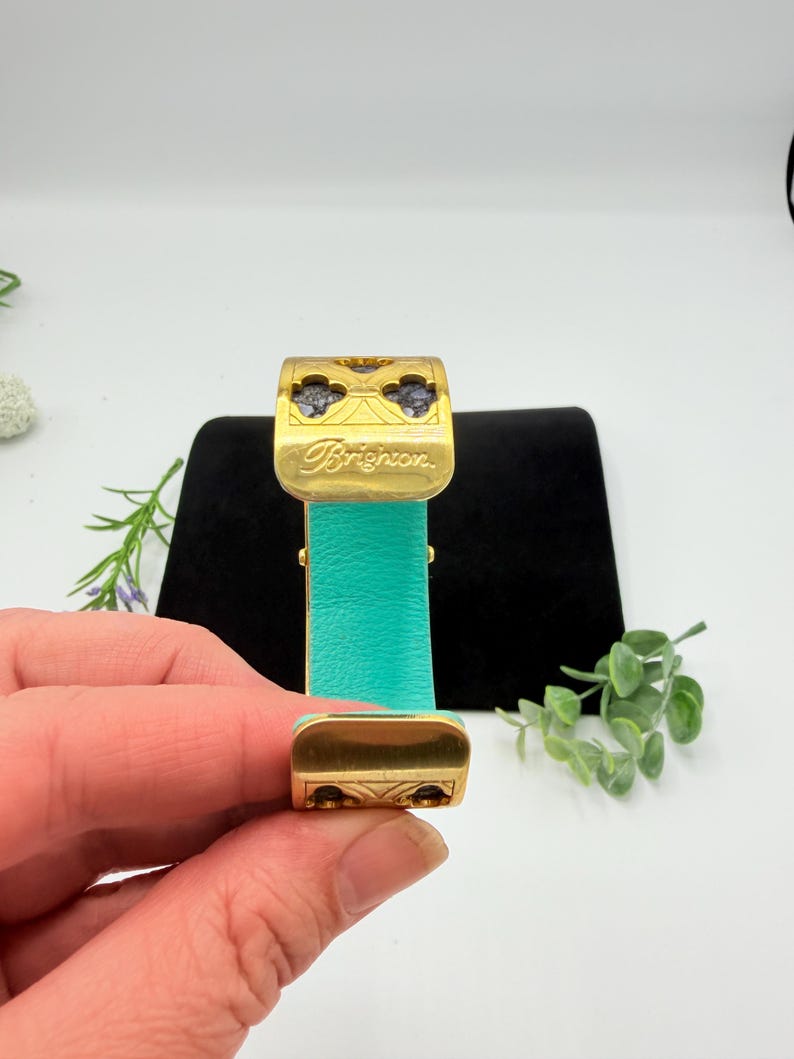 May include: A gold-tone metal cuff bracelet featuring a bright turquoise leather inlay. The top section displays a cutout clover pattern with dark stone accents and the word Brighton engraved in script. The design includes a hinged mechanism.