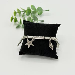 May include: A silver-toned charm bracelet featuring cylindrical beads with etched patterns and dangling charms shaped like a starfish, a seashell, and a small sea creature, all displayed on a small black velvet pillow.