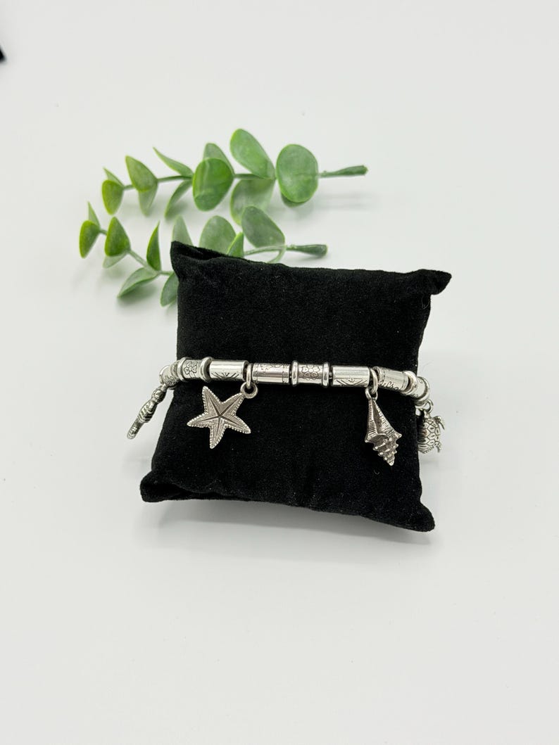 May include: A silver-toned charm bracelet featuring cylindrical beads with etched patterns and dangling charms shaped like a starfish, a seashell, and a small sea creature, all displayed on a small black velvet pillow.