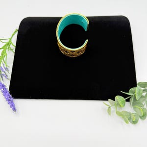 May include: A gold-toned cuff bracelet featuring an intricate openwork lattice pattern on the exterior and a smooth, vibrant turquoise-coloured interior lining. The bracelet has a C-shaped open design, allowing it to be slipped onto the wrist.
