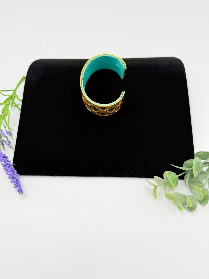 May include: A gold-toned cuff bracelet featuring an intricate openwork lattice pattern on the exterior and a smooth, vibrant turquoise-coloured interior lining. The bracelet has a C-shaped open design, allowing it to be slipped onto the wrist.