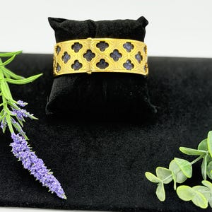 May include: A gold-toned cuff bracelet featuring a repeating quatrefoil cutout pattern. The metal frame overlays a textured, dark blue inlay that is visible through the geometric openings. The bracelet has a wide, flat band design.