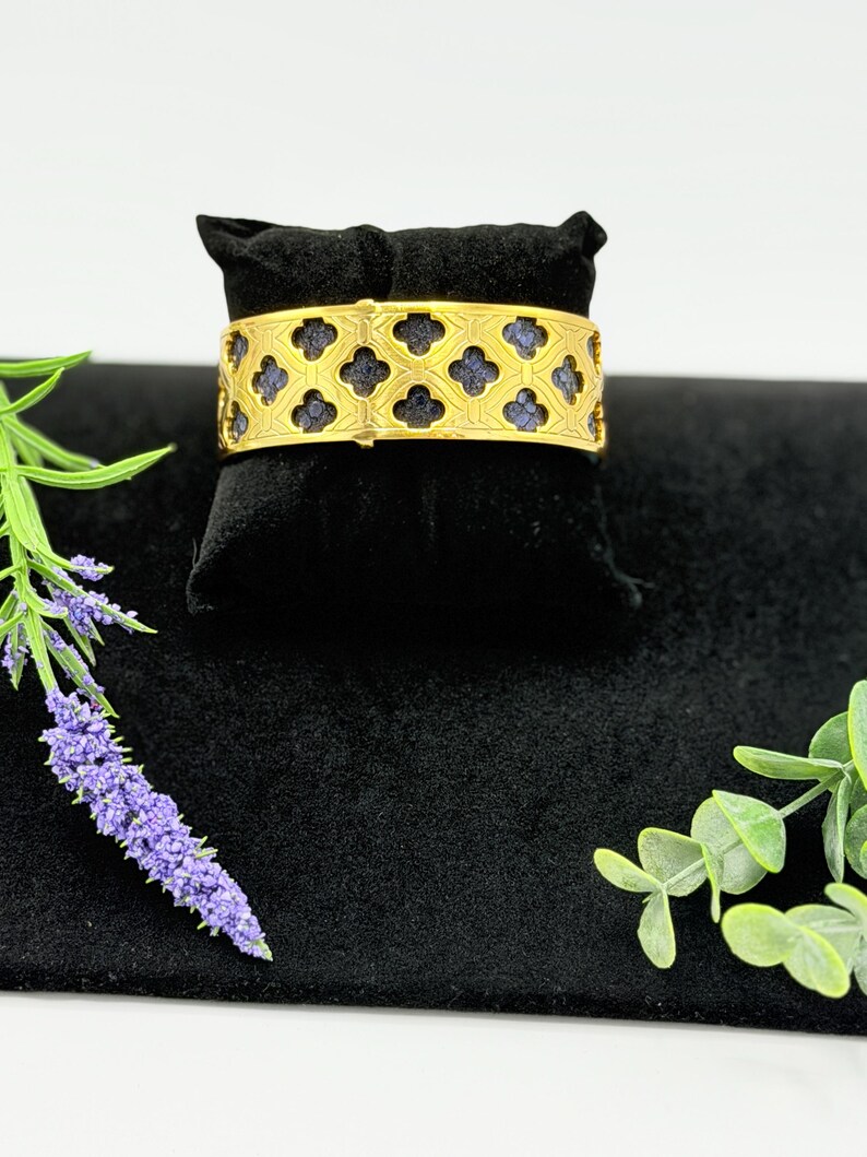 May include: A gold-toned cuff bracelet featuring a repeating quatrefoil cutout pattern. The metal frame overlays a textured, dark blue inlay that is visible through the geometric openings. The bracelet has a wide, flat band design.