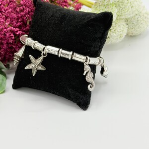 May include: A silver-toned charm bracelet featuring cylindrical beads with etched patterns and dangling charms shaped like a starfish, a seahorse, and small seashells. The bracelet is displayed on a small, square, black velvet pillow.
