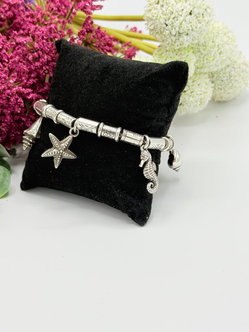 May include: A silver-toned charm bracelet featuring cylindrical beads with etched patterns and dangling charms shaped like a starfish, a seahorse, and small seashells. The bracelet is displayed on a small, square, black velvet pillow.