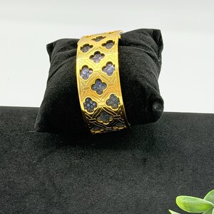 May include: A gold-toned cuff bracelet featuring a repeating quatrefoil cutout pattern. The metal frame has a polished finish, revealing a textured, dark blue inlay within each of the clover-shaped openings across the entire surface of the band.