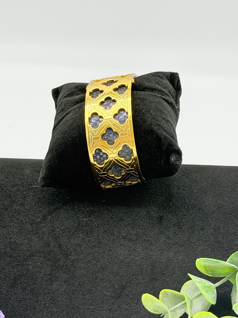 May include: A gold-toned cuff bracelet featuring a repeating quatrefoil cutout pattern. The metal frame has a polished finish, revealing a textured, dark blue inlay within each of the clover-shaped openings across the entire surface of the band.