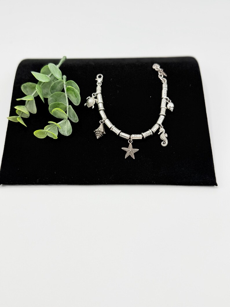 May include: A silver-toned charm bracelet featuring a series of cylindrical beads and multiple dangling charms, including a starfish, a sea turtle, a seahorse, and various seashells, arranged along a flexible metal chain with a lobster clasp.