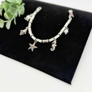 May include: A silver-toned charm bracelet featuring multiple nautical-themed pendants, including a starfish, a seahorse, and various seashells. The bracelet is constructed with cylindrical metal beads and a secure clasp mechanism.