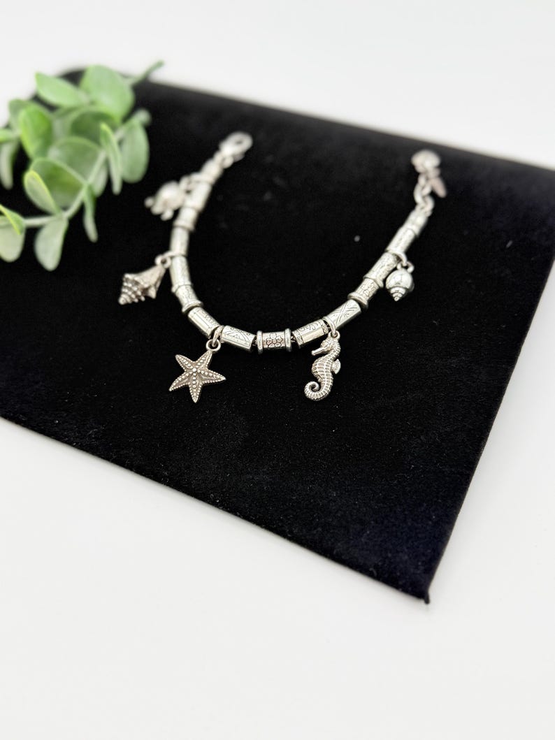 May include: A silver-toned charm bracelet featuring multiple nautical-themed pendants, including a starfish, a seahorse, and various seashells. The bracelet is constructed with cylindrical metal beads and a secure clasp mechanism.