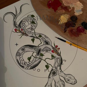 May include: A detailed black and white illustration of a two-headed snake, featuring dotwork shading, vines, leaves, and red mushrooms. The artwork is on a light background, with a wooden palette and paintbrushes visible.