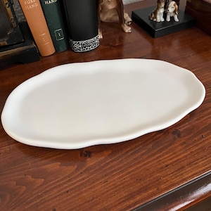 Peut inclure: Un plat de service en céramique blanc de forme organique. Le plat a une surface lisse et un bord légèrement irrégulier. Il est présenté sur une surface en bois foncé, suggérant un contexte décoratif ou de service.