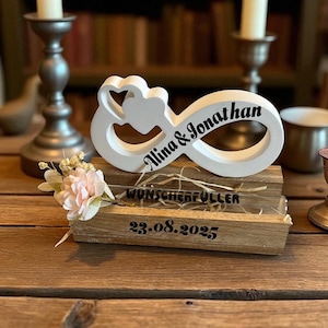 Peut inclure: Un symbole infini blanc avec deux cœurs, personnalisé avec les noms "Alina & Jonathan" en écriture noire. Il repose sur une base en bois avec le texte "WUNSCHERFÜLLER" et la date "23.08.2025". Un petit arrangement floral ajoute une touche délicate.