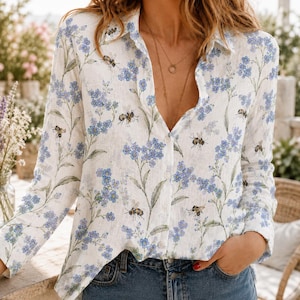 May include: A long-sleeved button-down shirt in white featuring a repeating pattern of small blue flowers and brown bees. The shirt has a classic collar, a front button placket, and a relaxed fit, showcasing a detailed botanical and insect print.