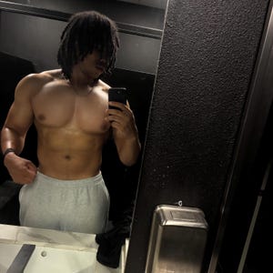 Op de afbeelding: Een persoon met dreadlocks wordt weerspiegeld in een spiegel, met een telefoon in de hand. De persoon draagt een grijze joggingbroek en een zilveren ketting. De achtergrond is een donkere, getextureerde muur en een roestvrijstalen armatuur.