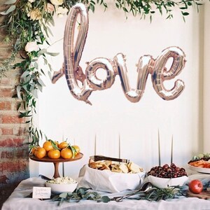 Puede incluir: Un globo de lámina de oro rosa que deletrea "love" cuelga frente a una pared de ladrillo decorada con vegetación. Una mesa está preparada con comida y bebida, incluyendo naranjas, pan y cerezas.