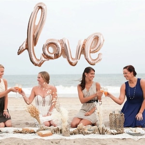 Puede incluir: Cuatro mujeres en una playa brindan con bebidas, con un gran globo "love" de oro rosa flotando sobre ellas. Están sentadas en una manta con objetos decorativos. El océano y el cielo están al fondo.