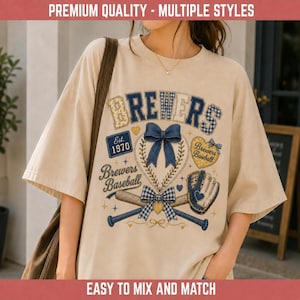 Puede incluir: Una camiseta beige de manga corta con un estampado gráfico que incluye el texto "BREWERS", "Est. 1970" y "Brewers Baseball". El diseño presenta un lazo azul estilo mezclilla, una pelota de béisbol, un guante, un bate y detalles en forma de corazón.