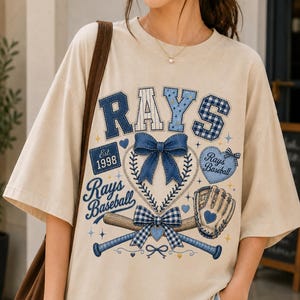 May include: A beige short-sleeve t-shirt featuring a graphic design with the text RAYS, Est. 1998, and Rays Baseball. The print includes a blue denim-textured bow, a baseball, a baseball glove with a heart, and two crossed baseball bats.