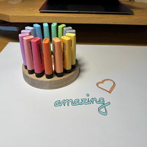 May include: A wooden pen holder with a circular design holds a collection of colorful highlighters. The highlighters are in shades of pink, orange, yellow, green, and blue. The word "amazing" is written in teal script with an orange heart.