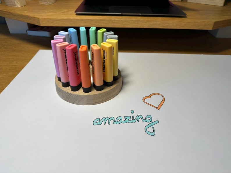 May include: A wooden pen holder with a circular design holds a collection of colorful highlighters. The highlighters are in shades of pink, orange, yellow, green, and blue. The word "amazing" is written in teal script with an orange heart.