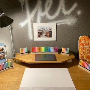 May include: A well-lit desk setup with a laptop, colorful markers, and a framed cityscape print. The desk features a wooden monitor stand and a white sheet of paper, with a black lamp and a decorative wooden board.