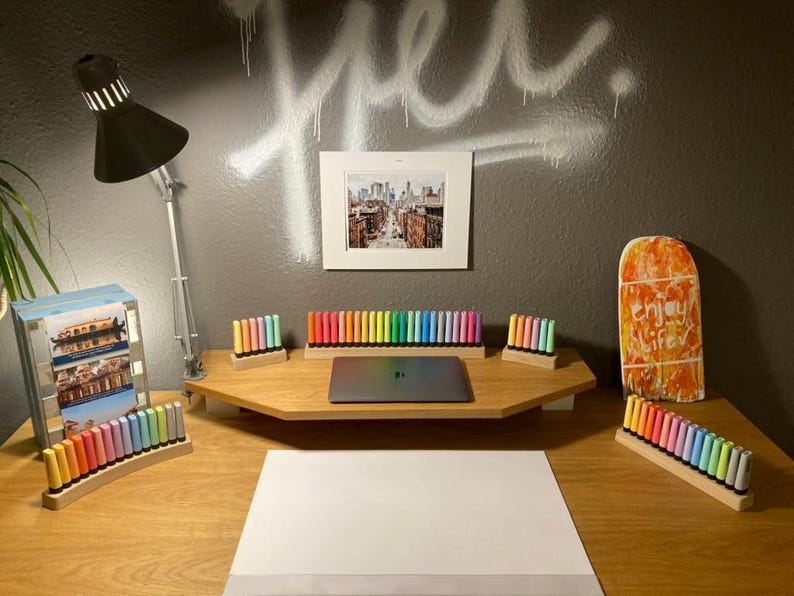 May include: A well-lit desk setup with a laptop, colorful markers, and a framed cityscape print. The desk features a wooden monitor stand and a white sheet of paper, with a black lamp and a decorative wooden board.