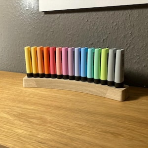 May include: A collection of colorful paint tubes arranged in a wooden holder. The tubes are in a rainbow of colors, including yellow, orange, pink, purple, blue, green, and gray. The holder is made of light-colored wood.