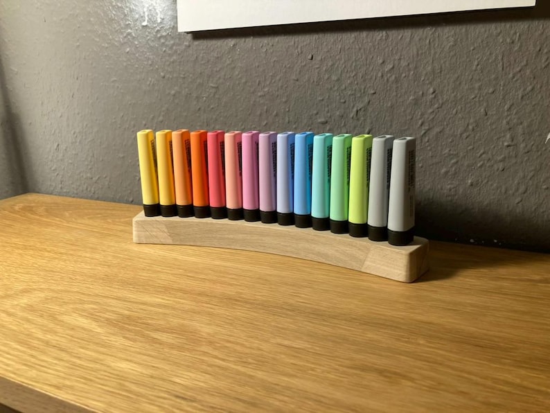 May include: A collection of colorful paint tubes arranged in a wooden holder. The tubes are in a rainbow of colors, including yellow, orange, pink, purple, blue, green, and gray. The holder is made of light-colored wood.
