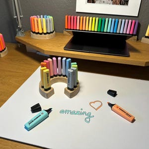 May include: A collection of colorful highlighters and pens arranged on a wooden desk. The image features various pen holders, including circular and arched designs. The word "amazing" is written on a white surface, along with a heart shape.