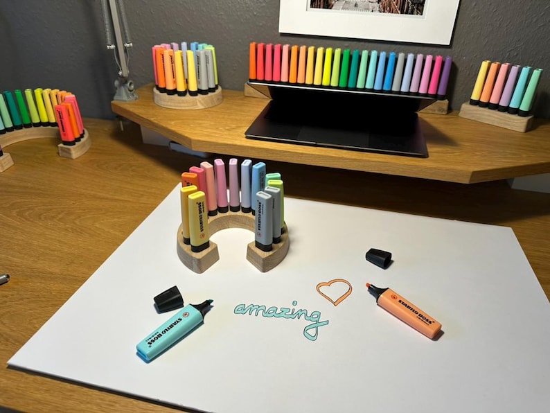 May include: A collection of colorful highlighters and pens arranged on a wooden desk. The image features various pen holders, including circular and arched designs. The word "amazing" is written on a white surface, along with a heart shape.