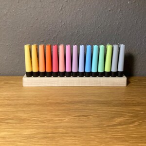 May include: A collection of colorful, cylindrical candles in a wooden holder. The candles are arranged in a row, showcasing a spectrum of colors from yellow to gray. Each candle has a black base, and the holder is made of light-colored wood.