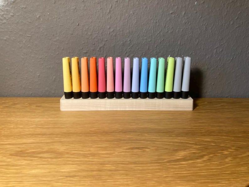 May include: A collection of colorful, cylindrical candles in a wooden holder. The candles are arranged in a row, showcasing a spectrum of colors from yellow to gray. Each candle has a black base, and the holder is made of light-colored wood.