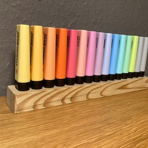 May include: A wooden organizer holds a collection of Stabilo Boss highlighters in various colors. The highlighters are arranged in a row, showcasing shades of yellow, orange, pink, purple, blue, green, and gray. The wooden organizer sits on a wooden surface.