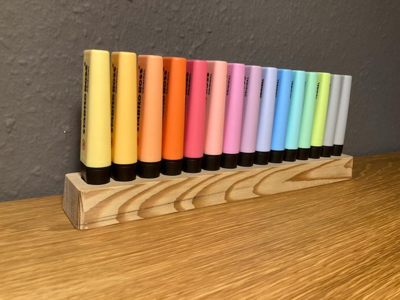 May include: A wooden organizer holds a collection of Stabilo Boss highlighters in various colors. The highlighters are arranged in a row, showcasing shades of yellow, orange, pink, purple, blue, green, and gray. The wooden organizer sits on a wooden surface.