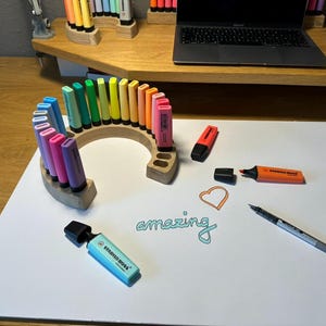 May include: A collection of colorful highlighters arranged in a wooden holder on a white surface. The word "amazing" is written in teal script. Other pens and a laptop are visible in the background.