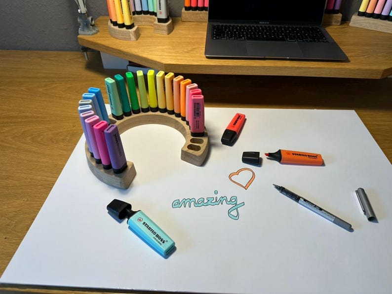 May include: A collection of colorful highlighters arranged in a wooden holder on a white surface. The word "amazing" is written in teal script. Other pens and a laptop are visible in the background.