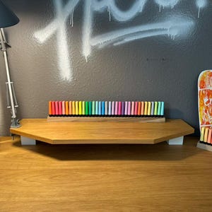 May include: A wooden desk with a variety of colorful markers arranged in a row. The markers are in a wooden holder. A desk lamp and a decorative skateboard are also visible.