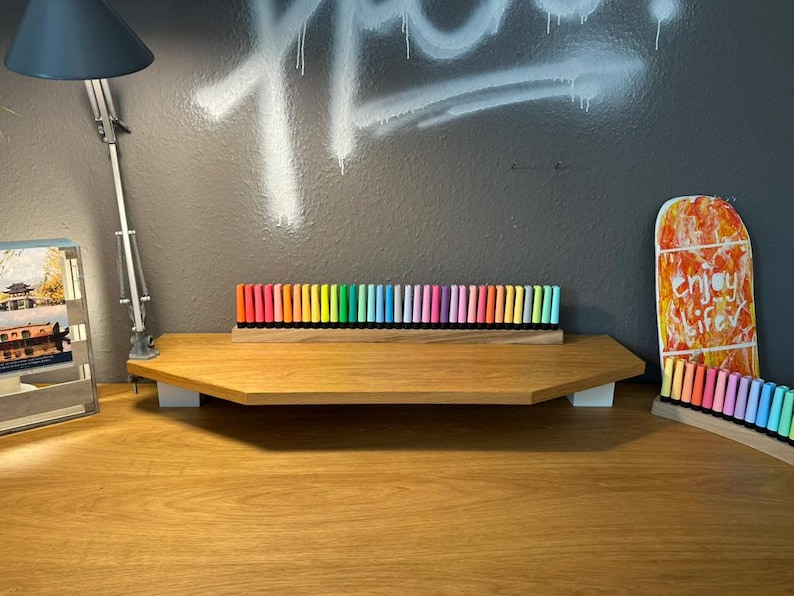 May include: A wooden desk with a variety of colorful markers arranged in a row. The markers are in a wooden holder. A desk lamp and a decorative skateboard are also visible.