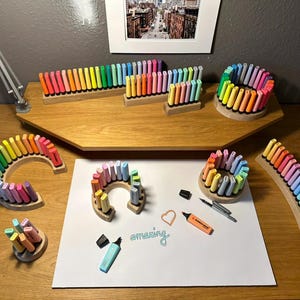 May include: A collection of colorful highlighters arranged in wooden holders. The highlighters are in various shapes, including straight lines, arcs, and circles. The word "amazing" is written on a white surface, along with other stationery items.