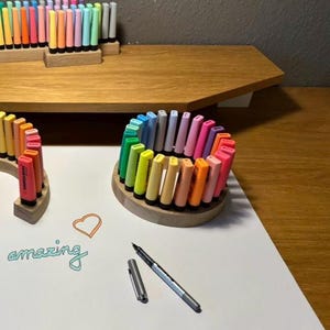 May include: A collection of colorful markers arranged in wooden holders. The markers are in various shades, including pink, orange, yellow, and green. A pen and the word "amazing" are also visible on the white surface. The markers are arranged in a circular pattern.