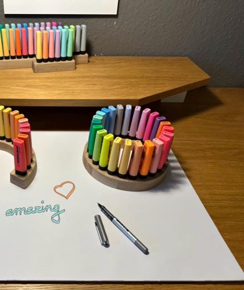 May include: A collection of colorful markers arranged in wooden holders. The markers are in various shades, including pink, orange, yellow, and green. A pen and the word "amazing" are also visible on the white surface. The markers are arranged in a circular pattern.