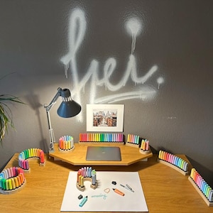 May include: A wooden desk with various colorful markers arranged in holders. A laptop, a framed picture, and a desk lamp are also on the desk. The wall behind the desk has white graffiti art.