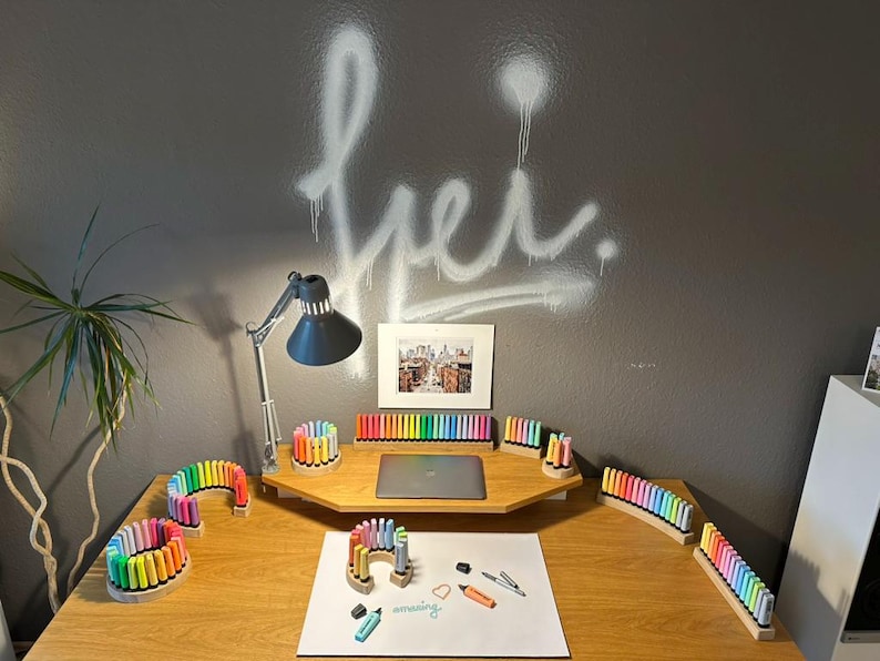 May include: A wooden desk with various colorful markers arranged in holders. A laptop, a framed picture, and a desk lamp are also on the desk. The wall behind the desk has white graffiti art.
