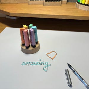 May include: A wooden pen holder with six pastel-colored highlighters. The word "amazing" is written in teal script on the white paper, along with an orange heart. A silver pen and cap are also on the paper.