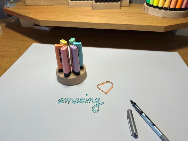 May include: A wooden pen holder with six pastel-colored highlighters. The word "amazing" is written in teal script on the white paper, along with an orange heart. A silver pen and cap are also on the paper.