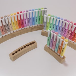 Può includere: Una collezione di evidenziatori colorati in supporti di legno. Gli evidenziatori sono disposti a ventaglio, mostrando uno spettro di colori. È presente anche un portapenne in legno separato.