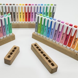 Può includere: Una collezione di evidenziatori colorati in varie tonalità, disposti in supporti di legno. Le penne hanno cappucci bianchi e sono esposte su una superficie bianca. Sono presenti anche diversi portapenne in legno.