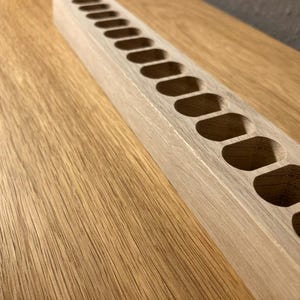 May include: A light-colored wooden organizer with a series of oval-shaped cutouts. The rectangular piece is positioned diagonally on a wooden surface, showcasing the natural wood grain and texture.