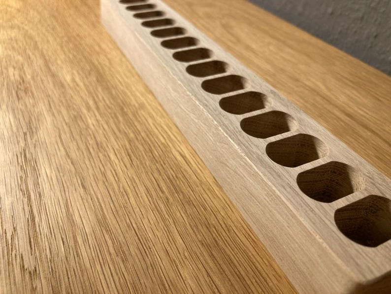 May include: A light-colored wooden organizer with a series of oval-shaped cutouts. The rectangular piece is positioned diagonally on a wooden surface, showcasing the natural wood grain and texture.